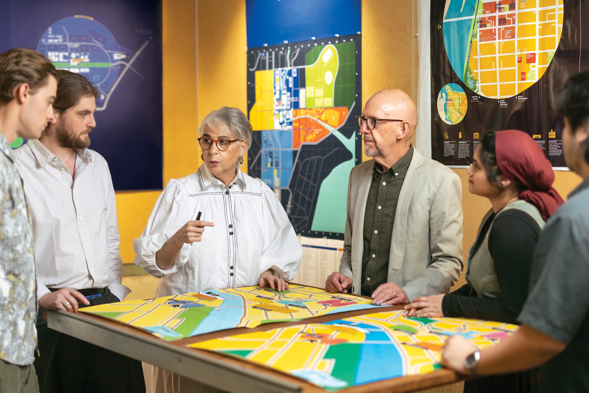 Five people gather around a large table covered with colourful urban planning maps, while additional map posters hang on the wall behind them. Despina Macris gestures with a pen as the group discusses the designs in a studio setting.