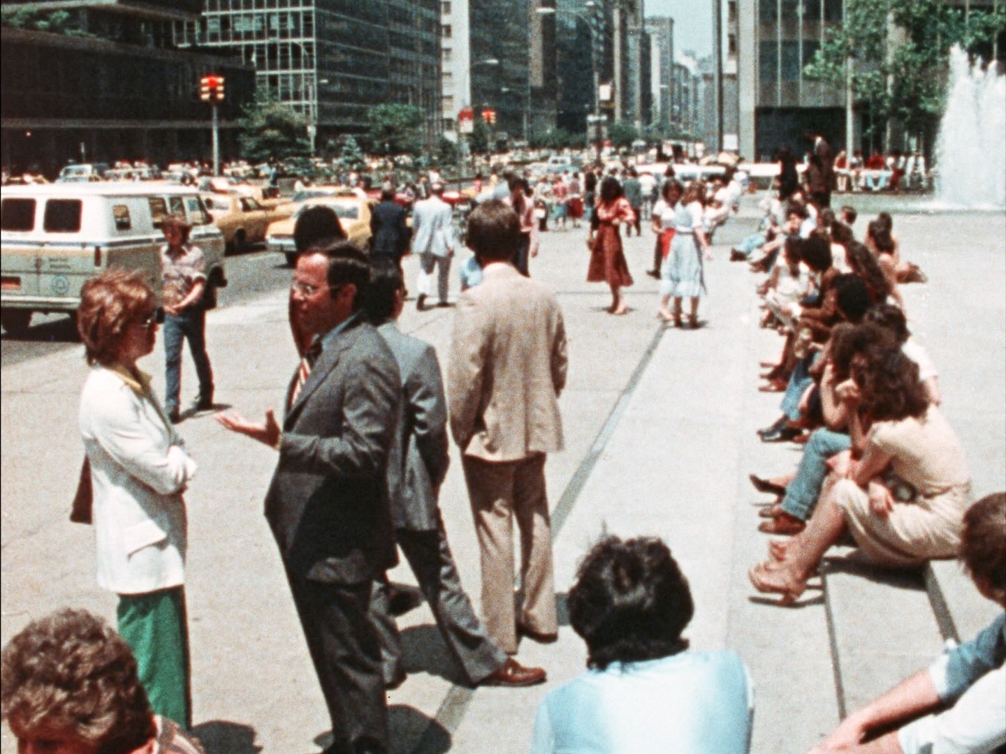 People standing and sitting on pavement next to a busy road, possibly in New York on a warm summer day