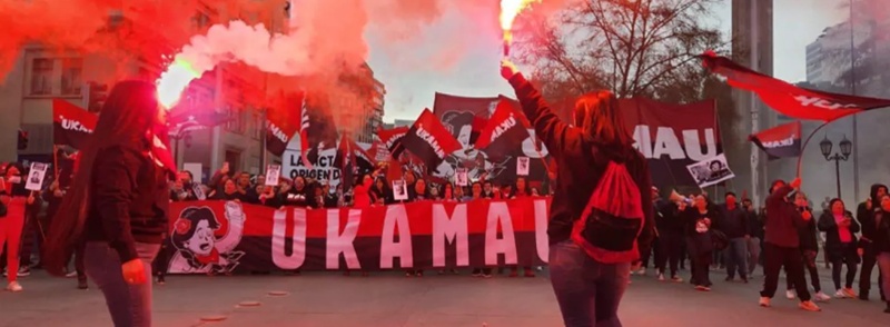 Two women in the foreground waving white / red flares above their heads, with a group of protestors standing behind a graphic banner in the background