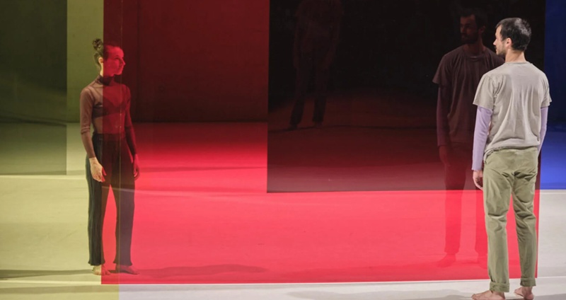Woman and man in casual clothing standing on a stage looking towards each other, with a large, red transparent screen seemingly between them.