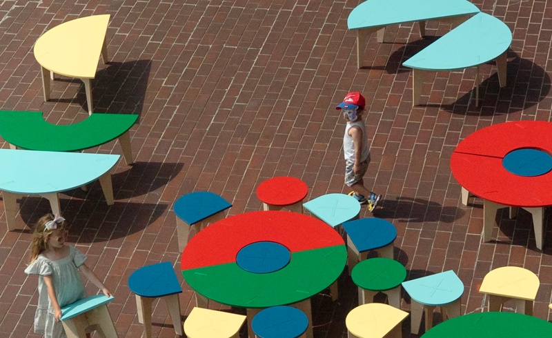 Foreground left, little girl wearing sunglasses and a summer dress carrying and moving a stool from one outside area to another (possibly to group it with others at a red, blue and green round table)