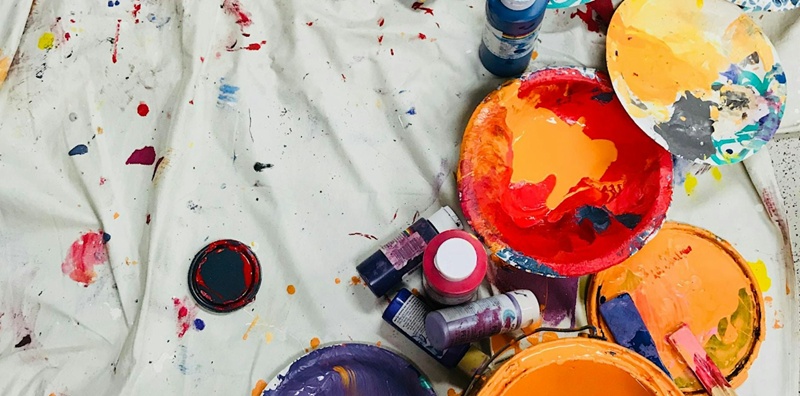 Various open paint tins and palettes on a white coloured cloth spattered with different coloured paints.