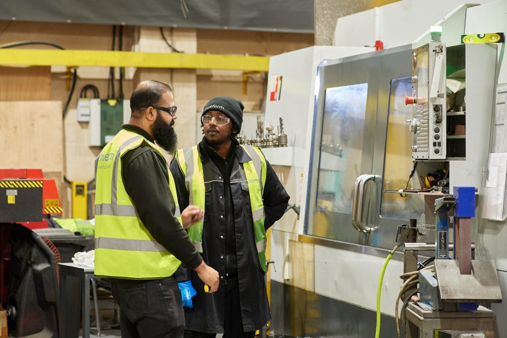 male work colleagues wearing black work clothes and high viz jackets, consulting in an indoor (factory?) setting.
