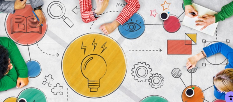 A group of people sit around a table covered with colourful illustrations representing creativity and teamwork. In the centre is a large yellow circle with a light‑bulb icon, surrounded by drawings such as gears, an eye, a magnifying glass, an open book, and arrows connecting the symbols. The individuals appear to be collaborating or brainstorming, with notebooks, coffee cups, and devices on the table.