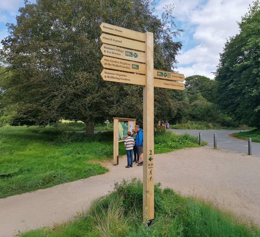 Timber wayfinding sign and map board at a park path junction in Stanmer Park, with visitors viewing the map and woodland surroundings.