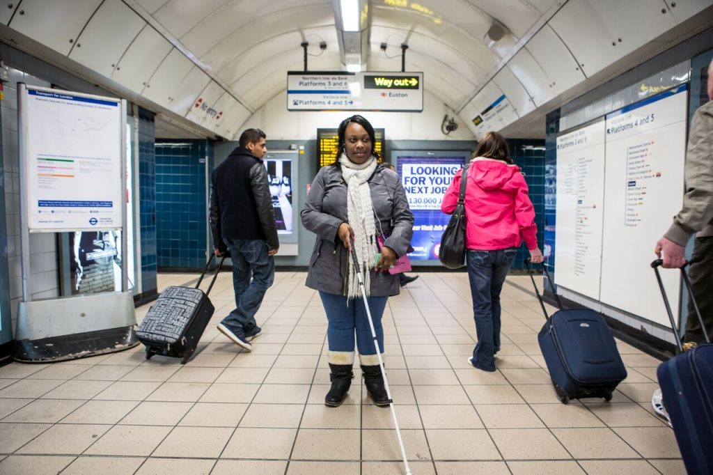 Sight-impaired woman using a white cane, in an Underground setting, with fellow travellers moving around her into the distance.