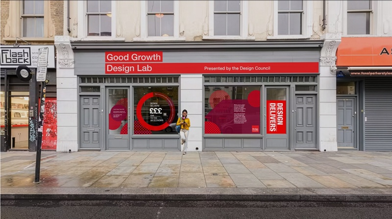 A single person standing outside two grey and red fronted units, named 'Good Growth Design Lab', part of a parade of shops, on their phone.