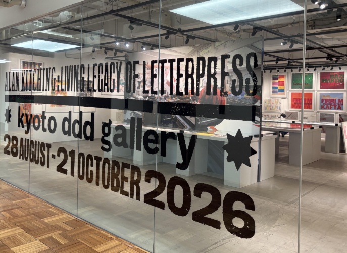 Entrance to an exhibition with large black lettering on the glass reading “Alan Kitching: A Living Legacy of Letterpress,” with gallery tables and colourful typographic artworks visible inside.