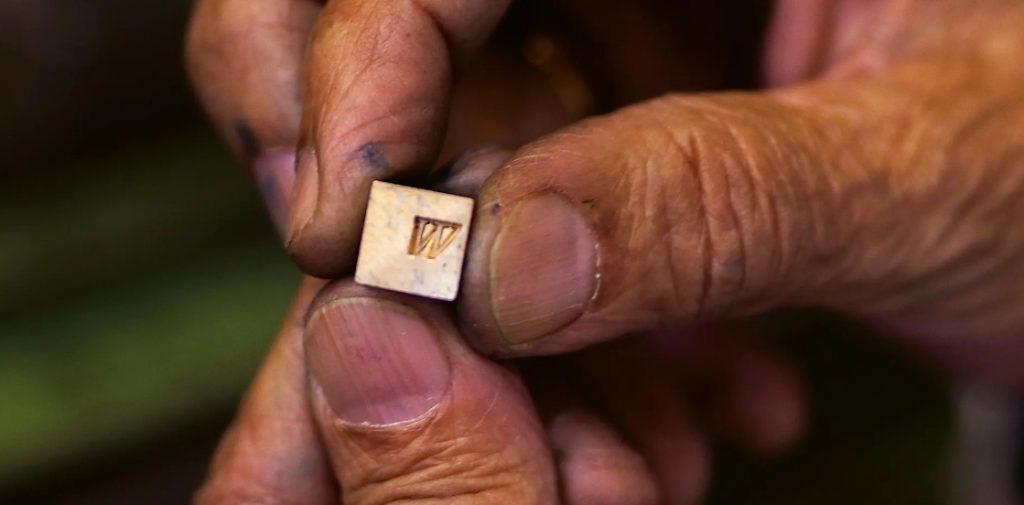 Close up of a grubby right hand holding a very small metal print block (letter 'W'), with left hand used for support beneath the print block.