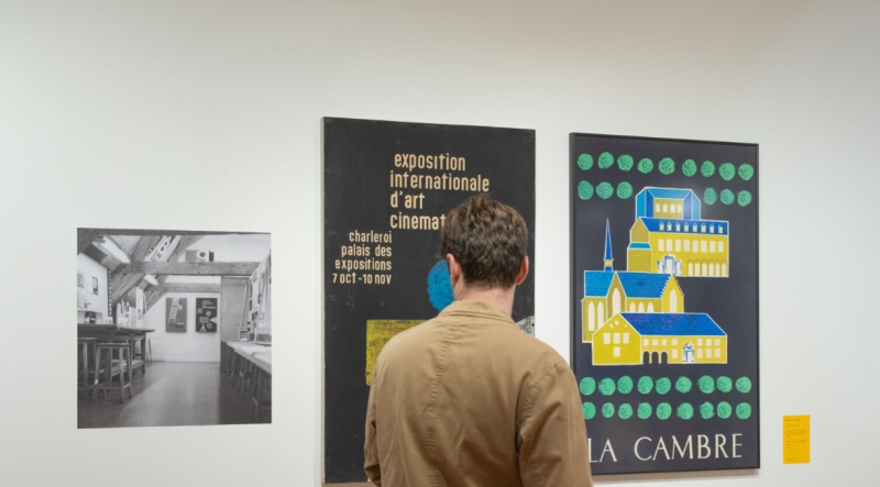 Visitor viewing graphic design posters in an exhibition gallery. Two large posters are mounted on a white wall alongside a black-and-white photograph of a workshop interior. The poster on the left features typography for an international cinema exhibition, while the poster on the right depicts a stylised architectural illustration with the words “La Cambre.” The gallery space is brightly lit, highlighting the exhibition's focus on graphic design and visual communication.
