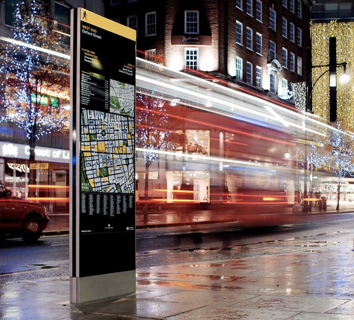 Blurred image of a red London bus passing down Oxford Street at night, with an Applied Information Group wayfinding totem featuring in the foreground (view is rainy, evening one)