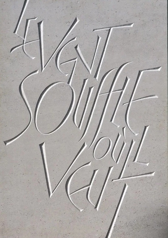 Excerpt: Stone surface with carved French lettering reading “LE VENT SOUFFLE OÙ IL VEUT,” engraved in a tall, stylised serif typeface.