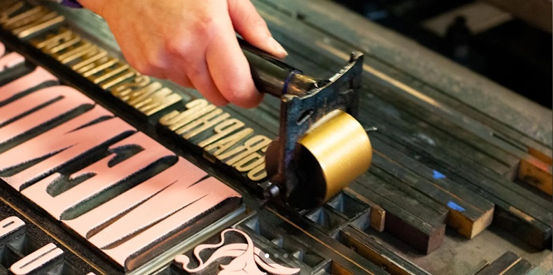 A hand rolls gold ink onto metal letterpress type, with some letters already inked pink.