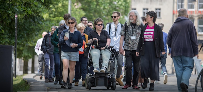 People (including two women in the foreground: one wearing a pink 'Living Streets' T-shirt, and the other in a wheelchair) walking as a group of circa 25, on a pavement in an urban environment.