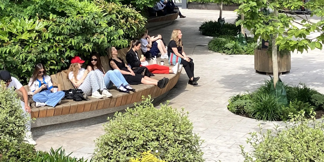 Girls and young women sitting on a curved bench in a park, some chatting togethers, others chilling, others eating and drinking, on a warm summer's day.