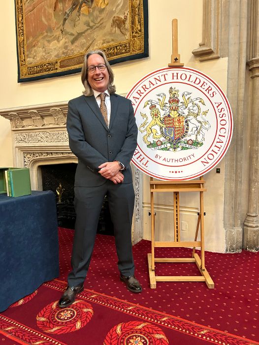 A smiling Mark Woolmer (Fitzpatrick Woolmer) at a Royal Warrant event, standing in an historic looking room, in front of an easel-supported 'The Warrant Holders Association' round plaque.