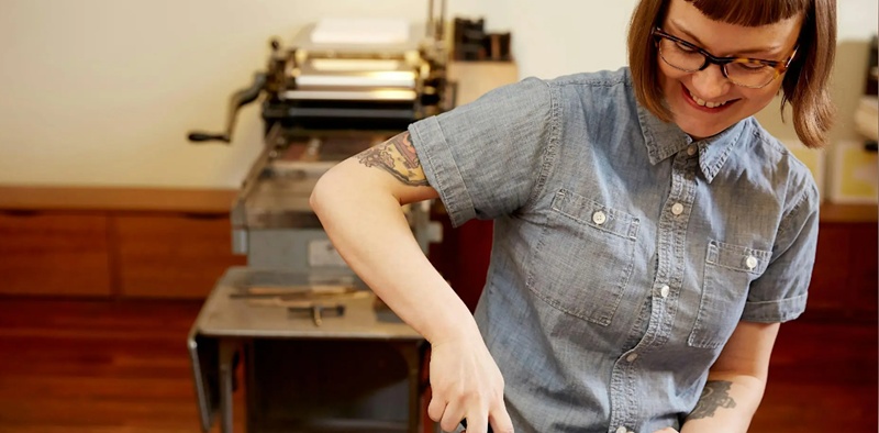 Photo of Erin Davis(with a smile on her face) demonstrating something in front of an old fashioned printing press.