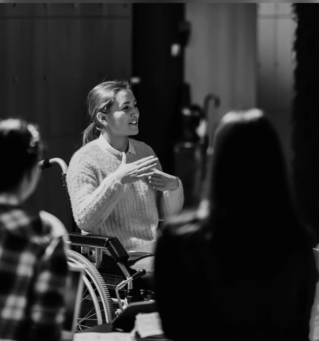 Black and white photo of a woman in a wheelchair talking to delegates at an unspecified event.