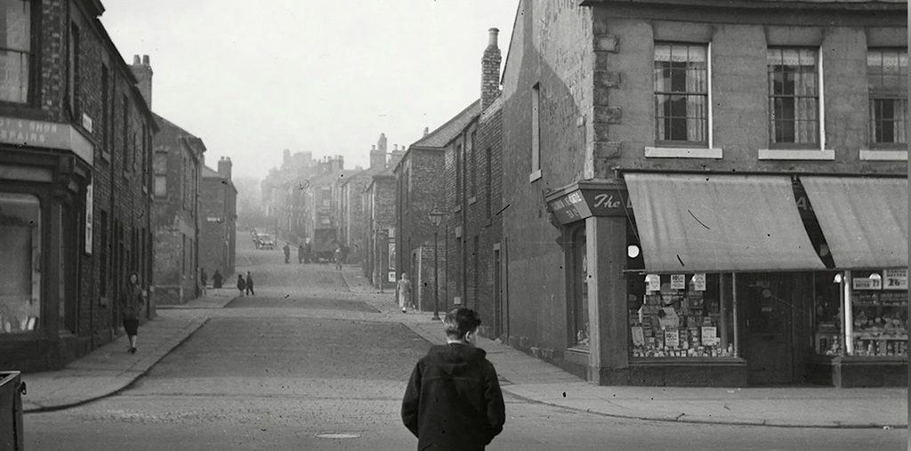 Historic black-and-white photograph of a quiet street lined with brick buildings and shopfronts. A lone pedestrian stands in the foreground while a few figures and vehicles appear in the distance. Traditional storefront signage and awnings reflect the character of a mid-century British city (in this case Newcastle).