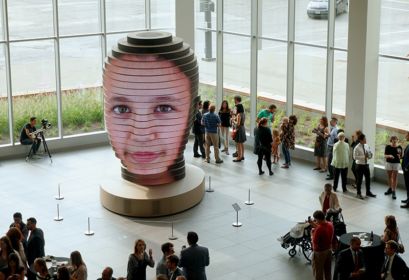 Human face shown on large modern 'head' feature in a spacious, windowed gallery. The feature 'head' is much bigger than the admiring visitors to the space.