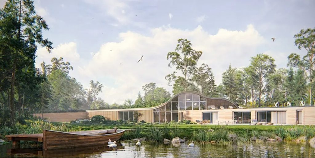 Modern building in the background, with lake in the foreground with small jetty and rowing boat. In a treed area on a bright summer's day.