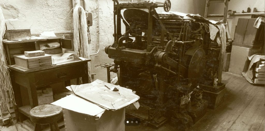 An old metal printing press sits in a small workshop, surrounded by a cluttered wooden desk with boxes, papers, and tools, creating a worn, historic print‑shop atmosphere.