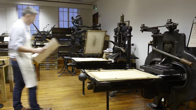 Blurred view of a man holding a large sheet of paper walking over to an old-fashioned printing press in situ at St. Bride Foundation.