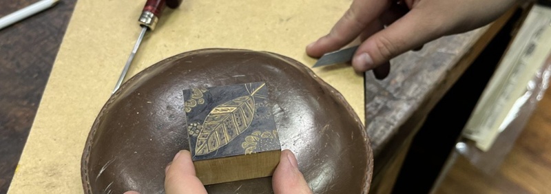 Hands holding a 'being-carved- wood block and carving tool, on a workbench.