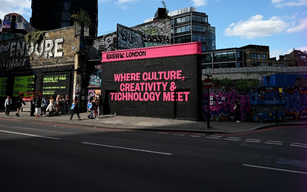 Promotional image showing SXSW London (Where Culture, Creativity and Technology Meet) painted in pink on a black wall in an urban setting next to a road