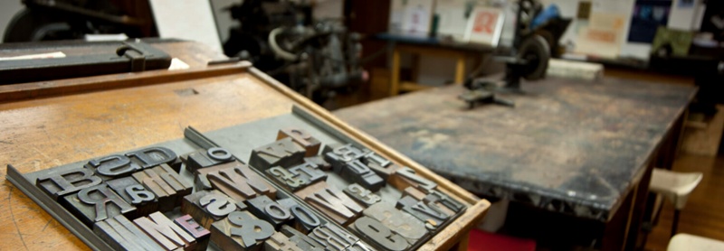 A tray of metal letterpress type in a workshop, with presses and printed materials visible in the background.