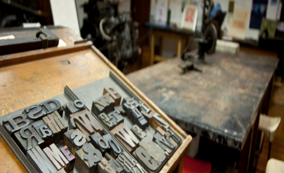 Close up of wooden alphabet letter type blocks, displayed on a semi-reclined wooden frame (in the foreground), juxtaposed against a print room background