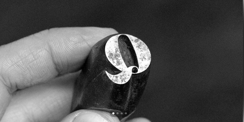 A close-up photograph of a hand holding a single piece of metal type. The metal block shows a serif uppercase “q” engraved on its end, slightly worn from use. The background is dark and out of focus, highlighting the detail and texture of the metal type.