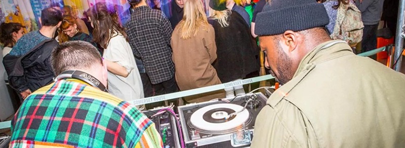 Two young men in the foreground looking at exhibits, with a group of young people chatting and enjoying themselves in the background.