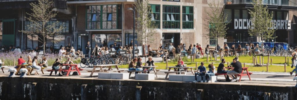 Promotional image for Utopian Hours 2026, due to take place in Rotterdam in late May of this year. Photo showing people sitting and standing around outside a modern building near water (a canal?) on a summer's day.