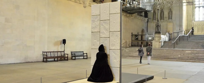 Figure in a long, black cloak standing staring at a brick wall installation in the middle of an unspecified historic building.