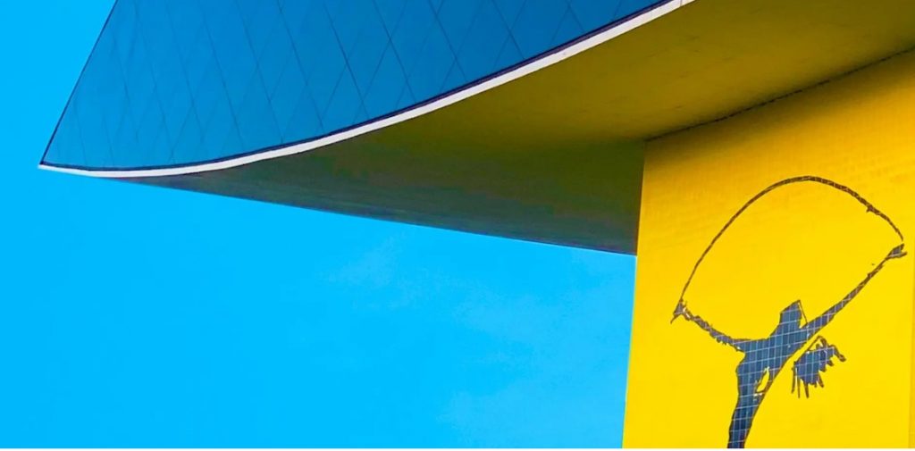 Blue and yellow part of a building (with blue outline of a girl with a skipping rope high above her head) against a clear, blue sky.